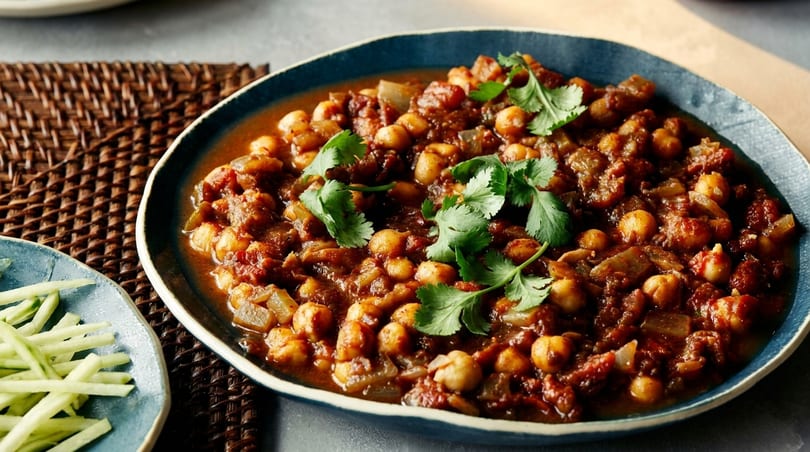 A blue bowl filled with chana masala, garnished with fresh cilantro, next to a plate of cucumber sticks.